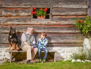 Ein Hund, ein älterer Mann und ein Bub sitzen auf einer Bank vor einer Holzhütte