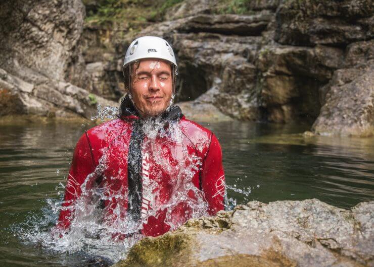 Mann mit Helm und Neoprenanzug taucht beim Canyoning in einer Schlucht aus dem Wildwasser auf
