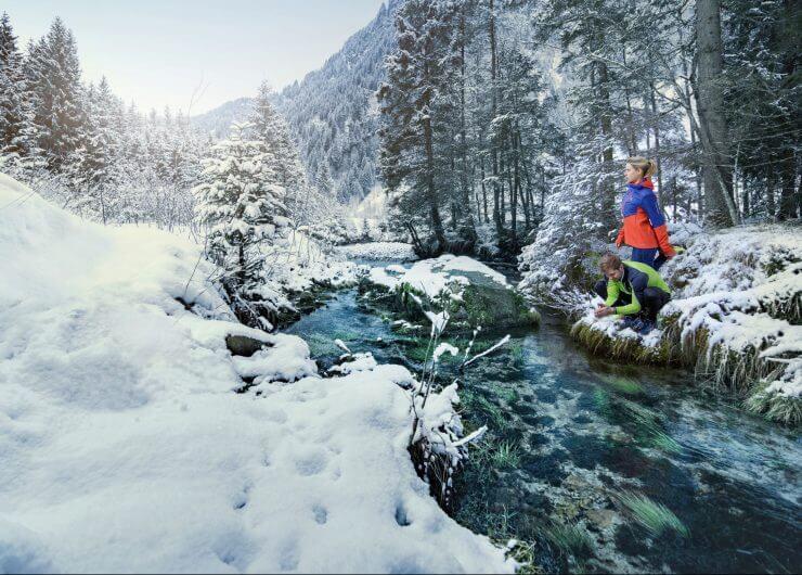 Zwei Läufer erfrischen sich am glasklaren Bach in einem verschneiten Wald