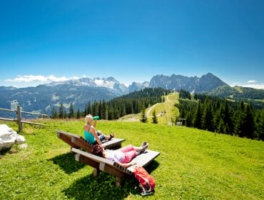 Wanderer entspannen auf zwei Holzliegen mit Blick auf die Bergwiesen und Gipfel