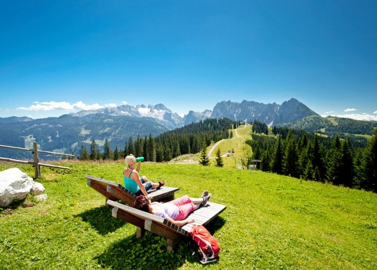 Wanderer entspannen auf zwei Holzliegen mit Blick auf die Bergwiesen und Gipfel