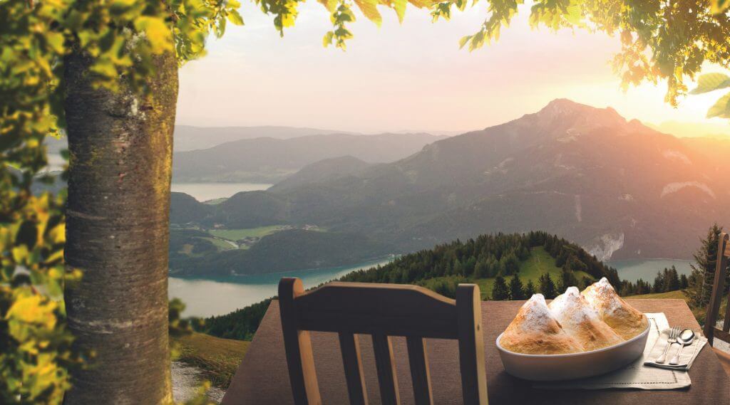 Salzburger Nockerl auf einem Tisch mit Sessel auf einem Berg, in der Ferne Seepanorama